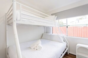 Ein Etagenbett mit weißer Matratze und Kissen, ein weißer Stuhl und ein Fenster mit Blick auf eine Ziegelmauer.