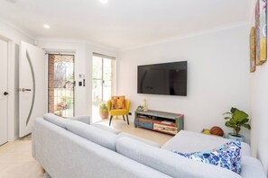 Een woonkamer met een grijze bank, een gele fauteuil, een aan de muur bevestigde televisie en een kleurrijk kastje.