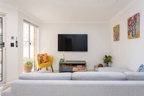 Ein Wohnzimmer mit grauem Sofa, einem gelben Sessel, einem Flachbildfernseher, einem bunten Couchtisch und zwei an der Wand hängenden Bildern.