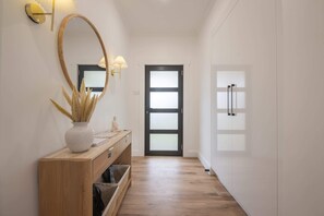 A hallway with a wooden floor, a white wall, a wooden console table with a vase and a sculpture, a mirror with a gold frame, and a black door with frosted glass panels. - Comfortable Family Home in Newstead (Newstead)