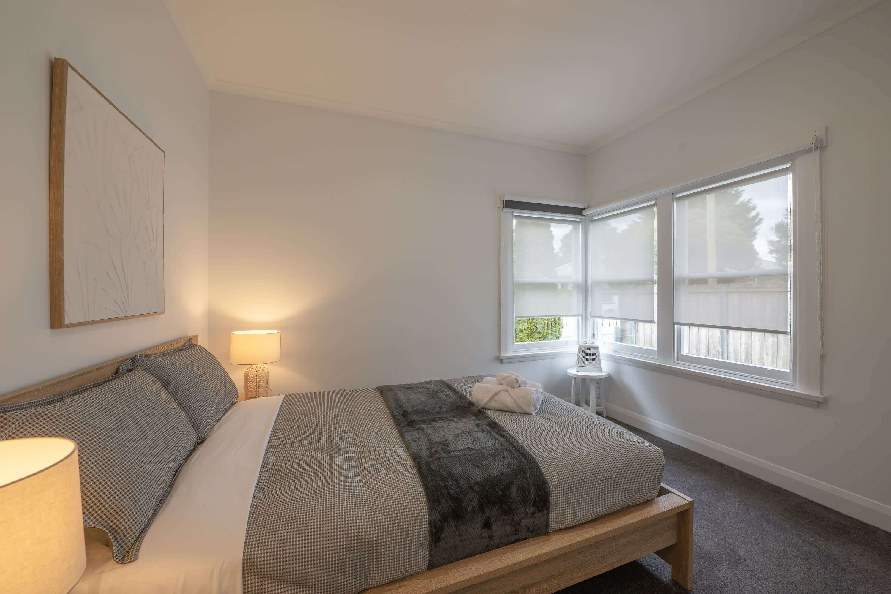A bedroom with a bed, a bedside lamp, a window with blinds, and a small table with a fan.