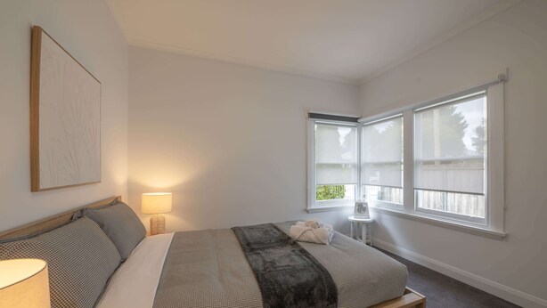 A bedroom with a bed, a bedside lamp, a window with blinds, and a small table with a fan. - Comfortable Family Home in Newstead (Newstead)