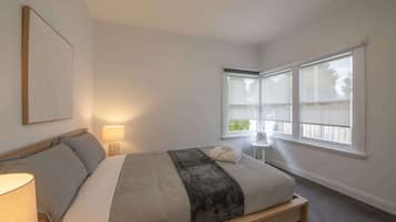 A bedroom with a bed, a bedside lamp, a window with blinds, and a small table with a fan.