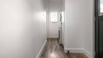 A narrow hallway with a wooden floor, a white door at the end, and a window with a view of the outside.