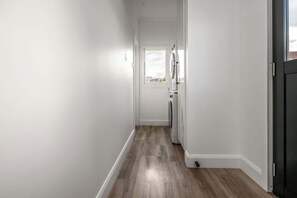 Um corredor estreito com piso de madeira, uma porta branca no final e uma janela com vista para o exterior.