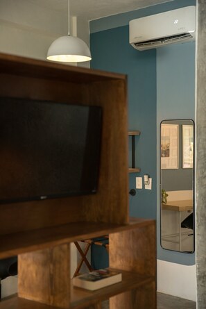Een kamer met een televisie op een houten standaard, een witte hanglamp, een blauwe muur, een airconditioner, een spiegel en een klein plankje met een boek.