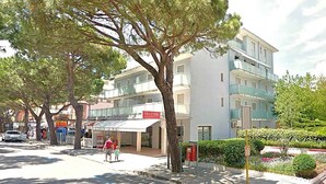 A street with a row of trees, a building with a sign that reads 'FREESB', and a sidewalk with a blue line marking. - Holiday apartment mit Klimaanlage und Balkon (Jesolo)