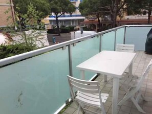 An outdoor patio with a white table and chairs, a glass railing, and a view of the street and buildings in the background. - Holiday apartment mit Klimaanlage und Balkon (Jesolo)