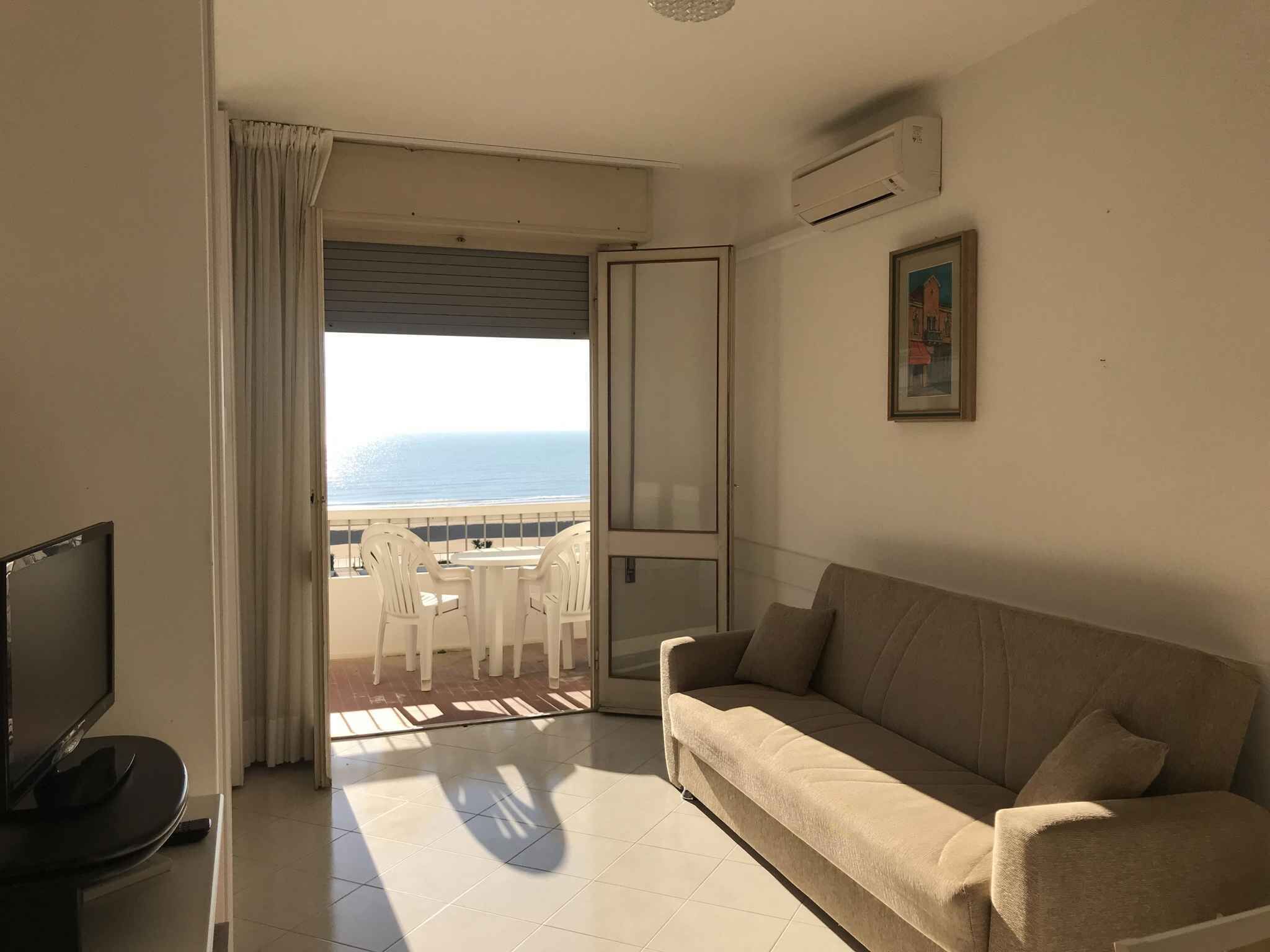 A room with a sofa, a TV, a balcony with a view of the sea, and an air conditioning unit.