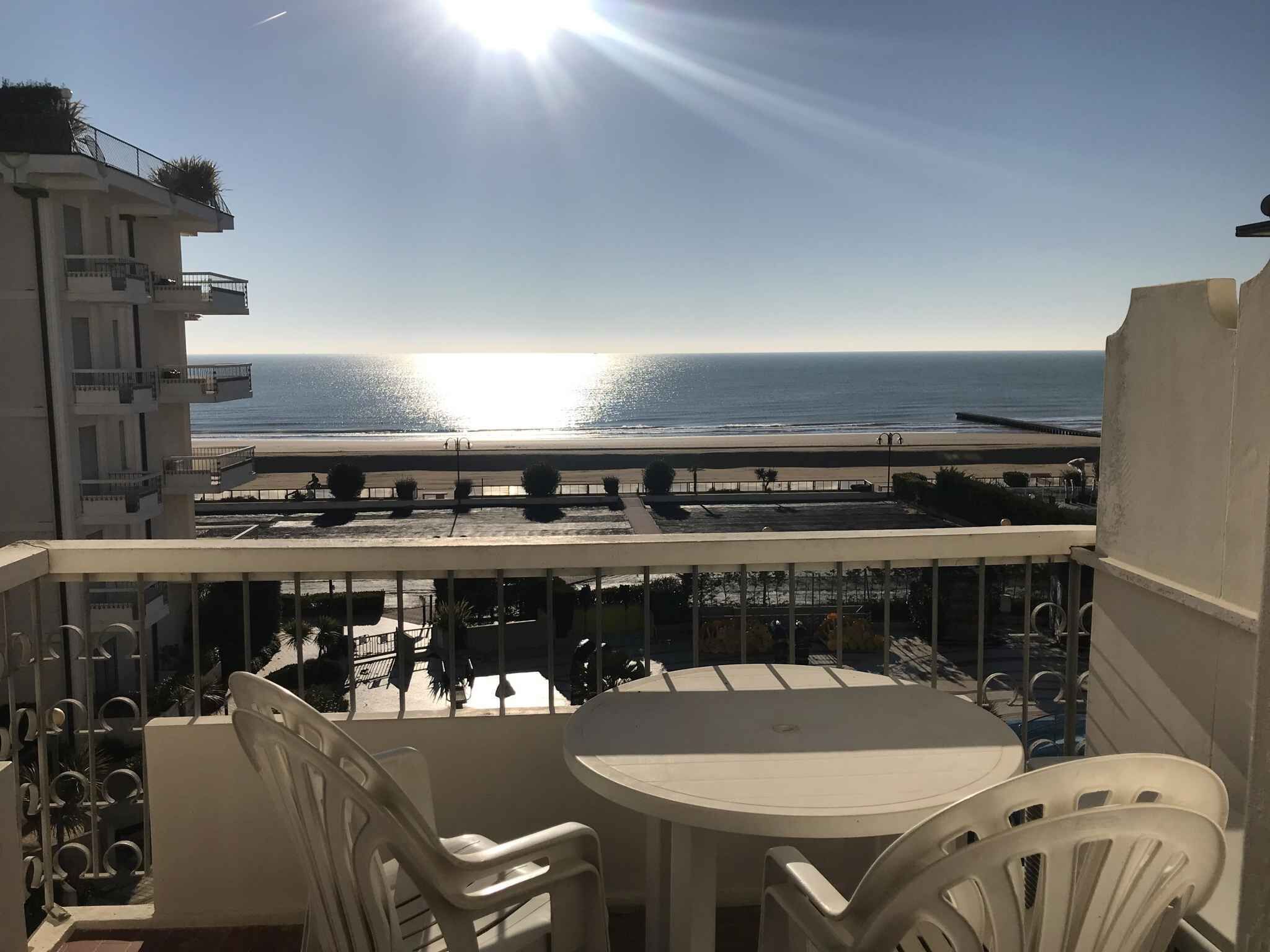 A balcony with a view of the beach and ocean, a round table, and white chairs.