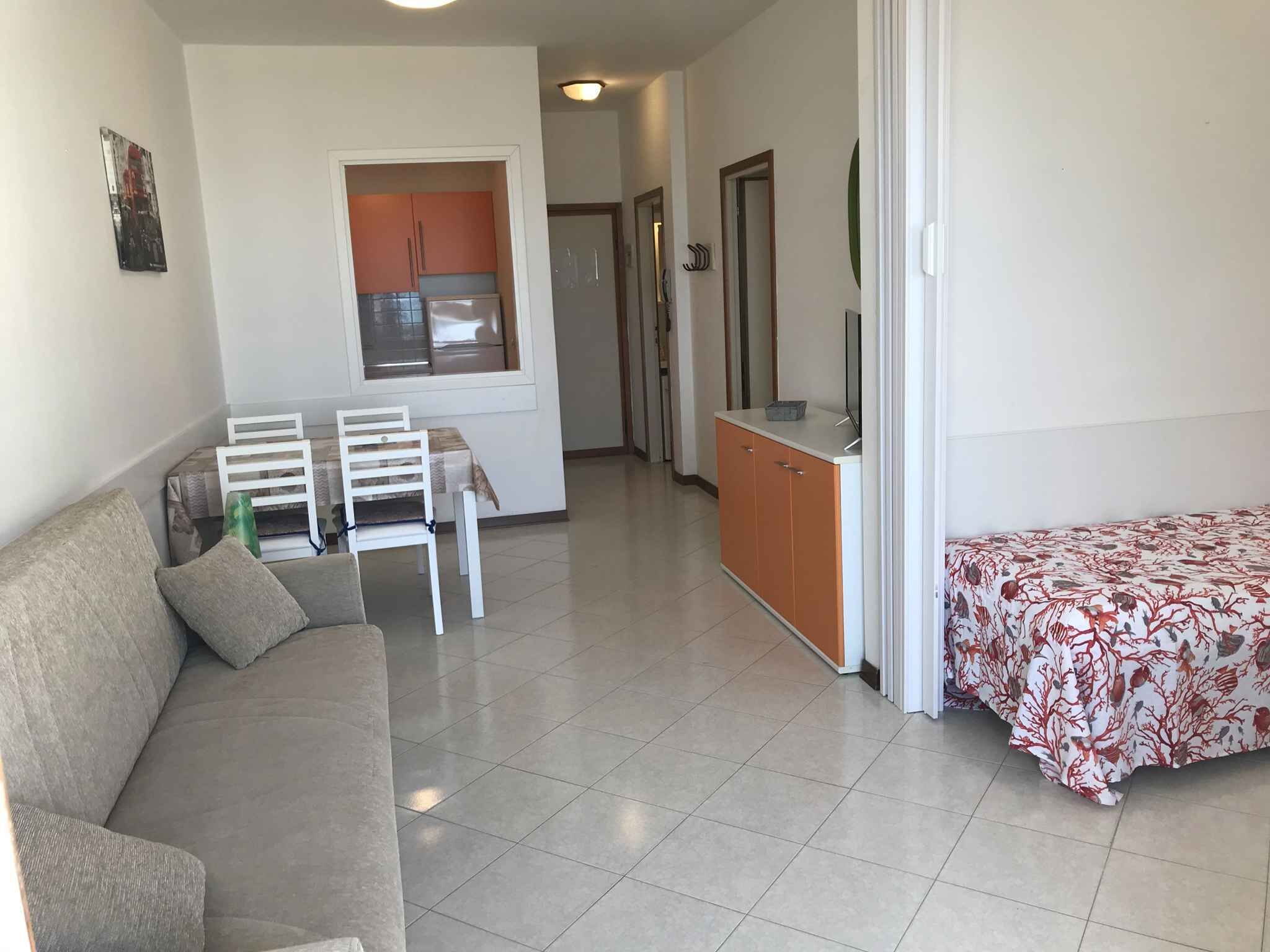 A living room with a sofa, a dining table with chairs, a kitchenette with cabinets, and a bed with a patterned bedspread.