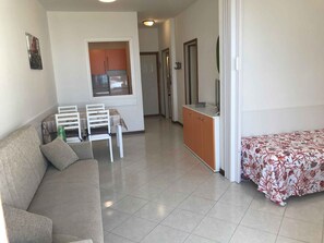 Ein Wohnzimmer mit Sofa, einem Esstisch mit Stühlen, einer Küchenzeile mit Schränken und einem Bett mit geblümtem Überwurf.