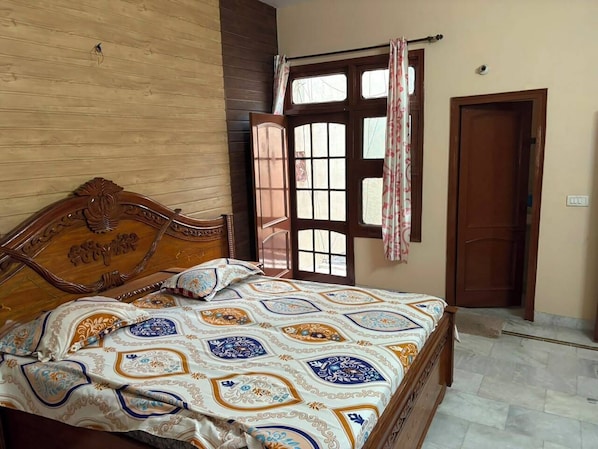 A bedroom with a wooden headboard, a patterned bedspread, a window with curtains, and a door.