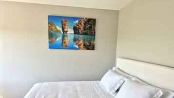 A hotel room with a large bed, white bedding, and a painting of a coastal landscape on the wall.