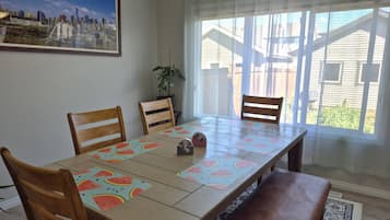 A dining room with a long table, chairs, a cityscape painting, a plant, and a view through sheer curtains.