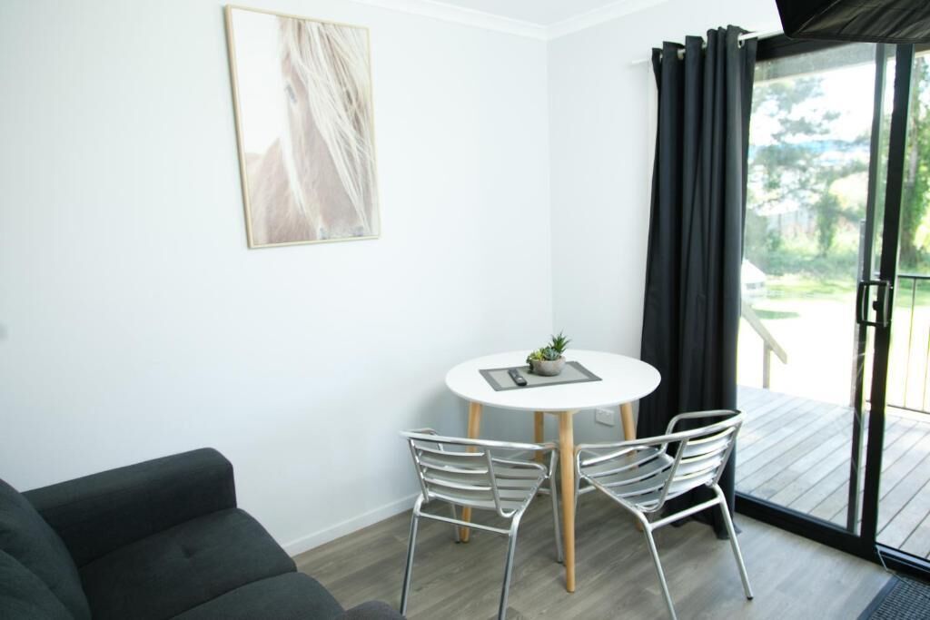 Una habitación con pared blanca, una mesa redonda con dos sillas, un sofá de color oscuro y un ventanal corredero que da a un espacio exterior.