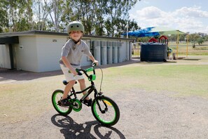 Un niño en bicicleta, con estructura verde, parado sobre una superficie pavimentada con un edificio y árboles al fondo.