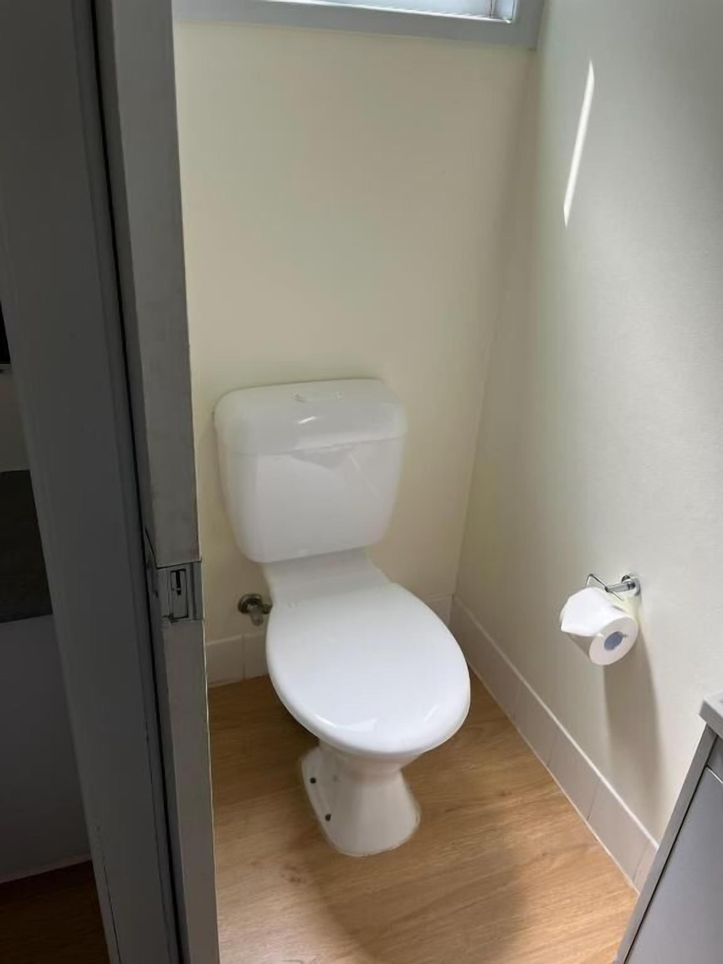 A small, compact bathroom with a white toilet, a wooden floor, and a wall-mounted toilet paper holder.