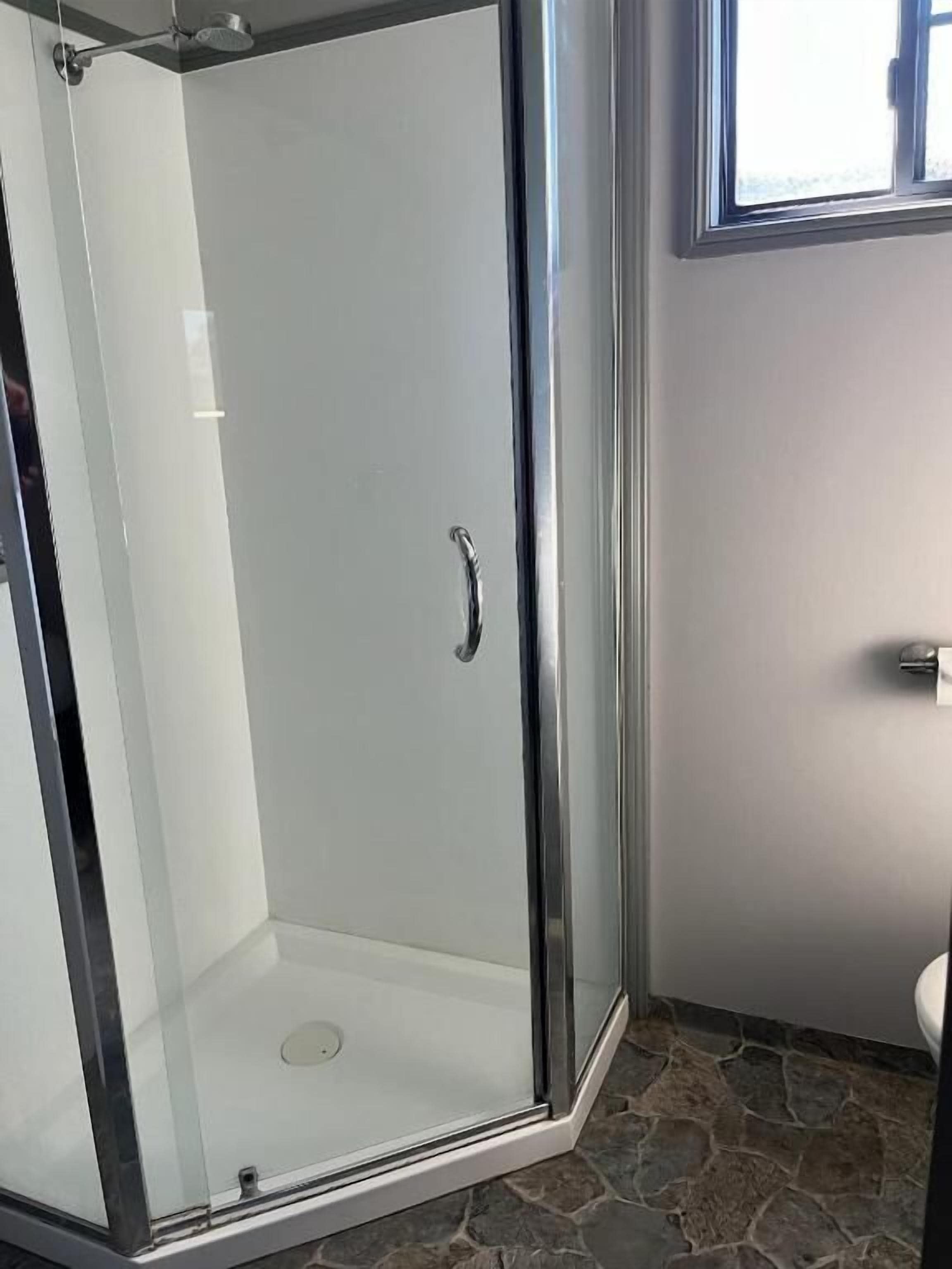 A glass-enclosed shower area with a door handle, adjacent to a toilet, in a bathroom with stone flooring.