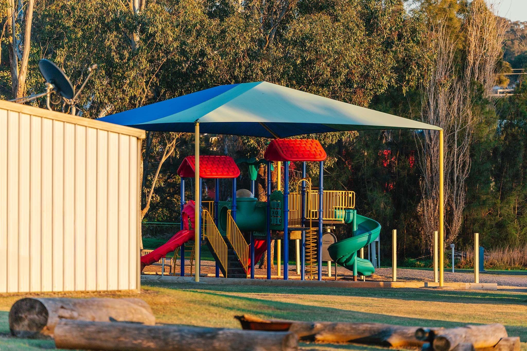 Un parque infantil con toboganes y estructuras para escalar, rodeado de árboles y un refugio cubierto.