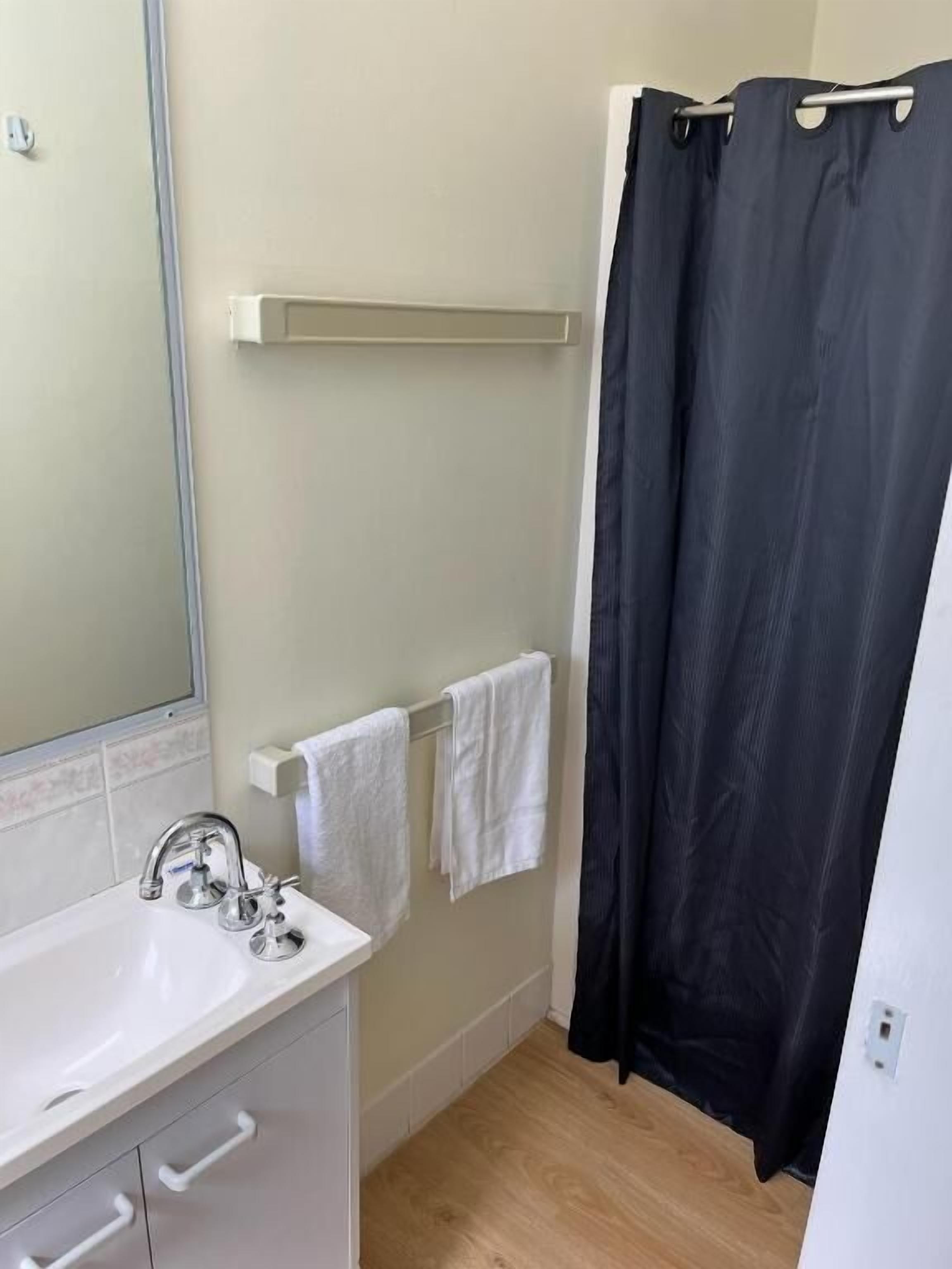 Un baño con un lavamanos blanco, un espejo, un perchero con dos toallas, una cortina de ducha negra y un suelo de madera.