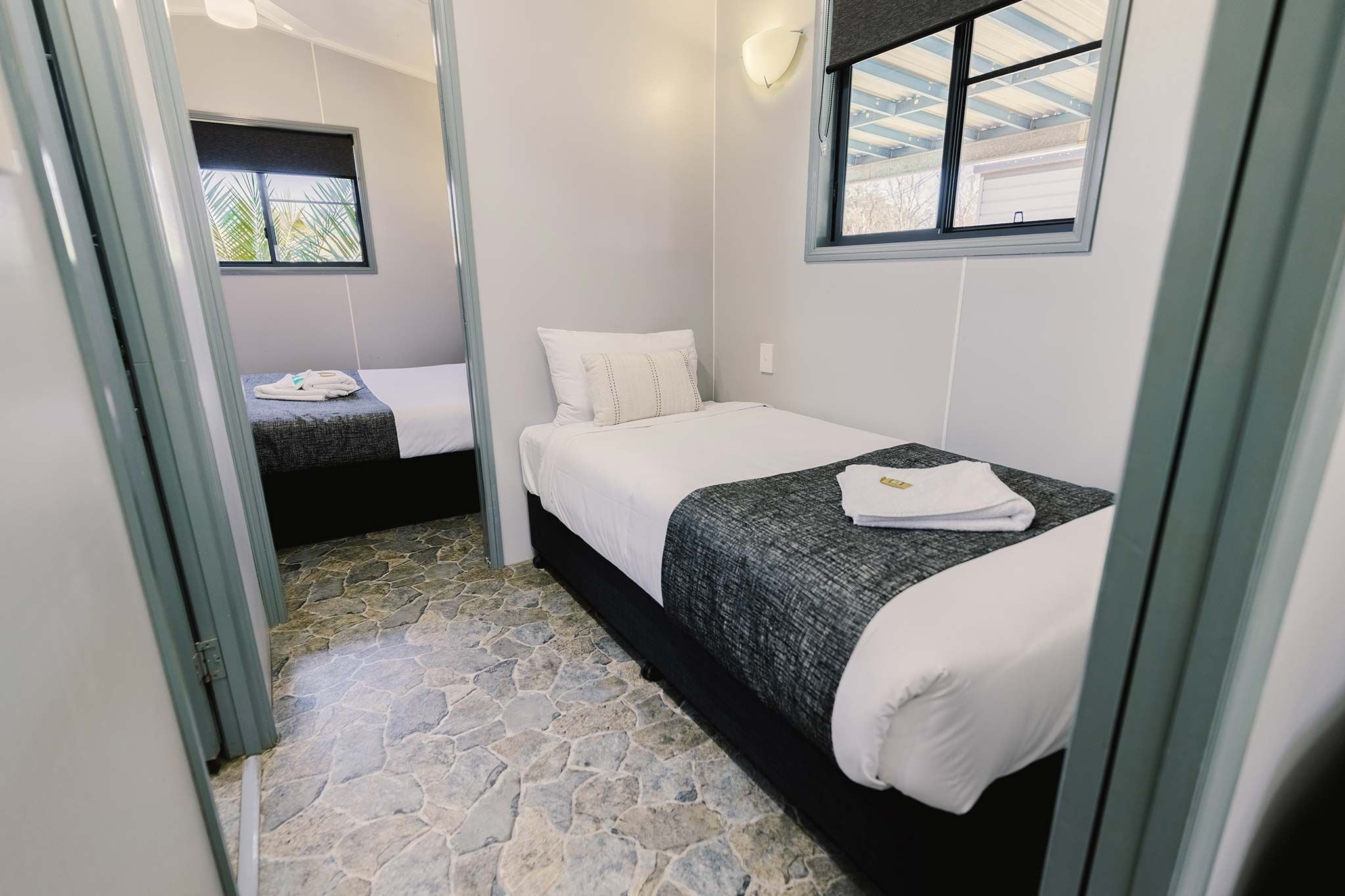 A small, compact room with two beds, a stone floor, and a window with a view of a garage.