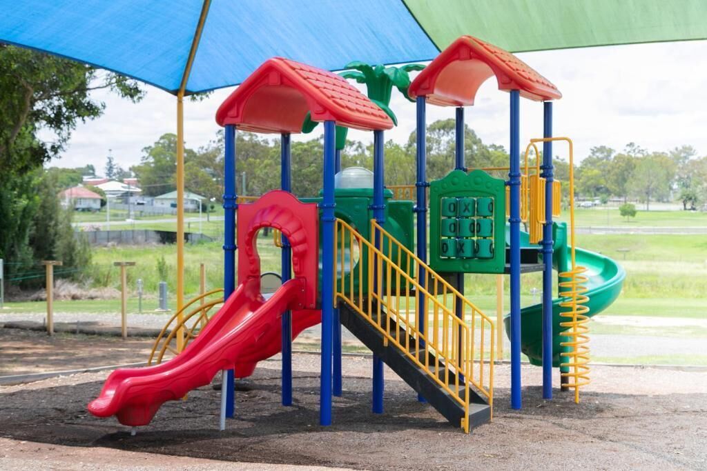 Um parquinho com um escorregador vermelho, estruturas de recreação amarelas e azuis, e uma área de escalada verde.