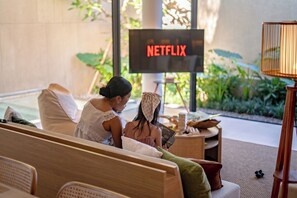 Una habitación con una televisión de pantalla plana grande mostrando 'NETFLIX' en letras rojas, un sofá, una mesita de noche con una lámpara y vistas a zonas verdes al exterior.