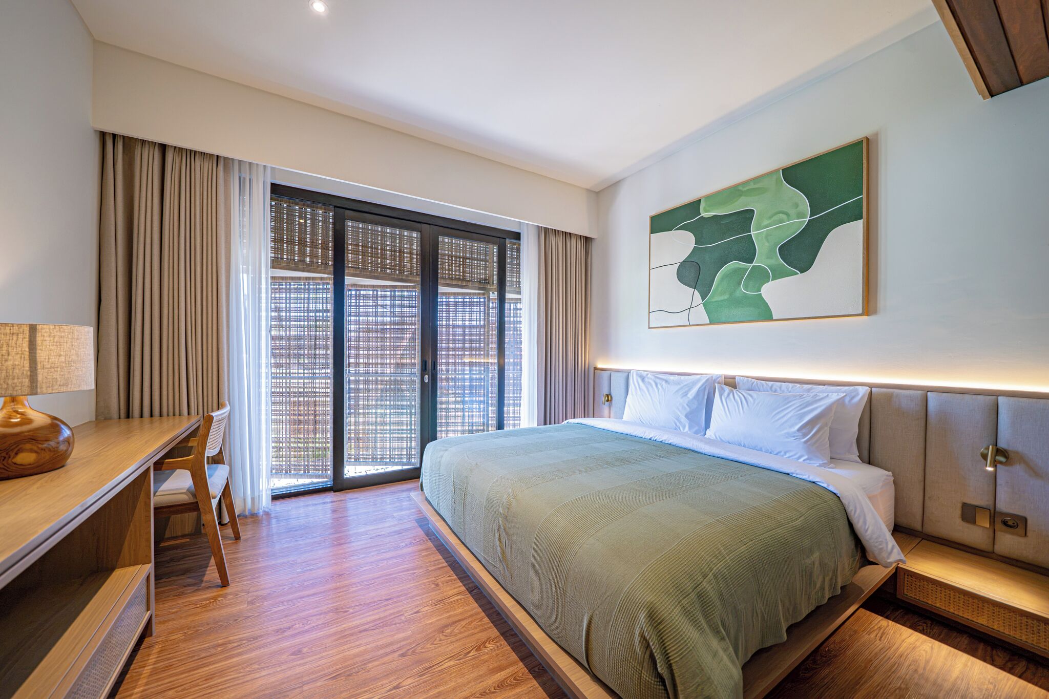Habitación de hotel con cama, escritorio, silla, ventana grande con cortinas y una obra de arte colgada en la pared.