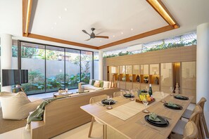 Un comedor moderno con una larga mesa de madera dispuesta para una comida, rodeada de asientos cómodos y vistas a la vegetación exterior.