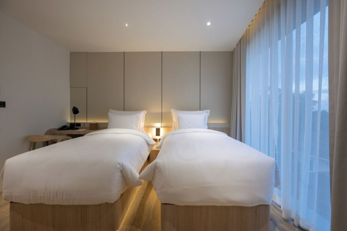 A modern hotel room with two beds, a nightstand, a lamp, and a large window with sheer curtains.