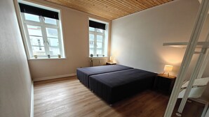 A bedroom with a wooden floor, two beds, a window with a view, and a nightstand with a lamp.