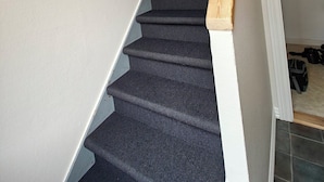 A staircase with dark carpeted steps and a wooden handrail, leading to a room with a door and a suitcase on the floor.