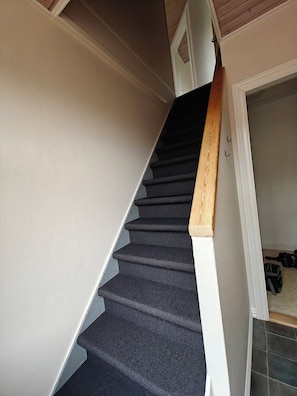 Un escalier avec des marches recouvertes d’une moquette sombre et une rampe en bois, menant à une pièce où se trouvent une porte et une valise posée sur le sol.