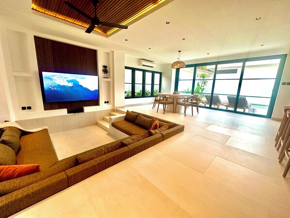 A modern living room with a large flat-screen TV, a sectional sofa, a dining area with a table and chairs, and large windows offering a view of the outdoors.
