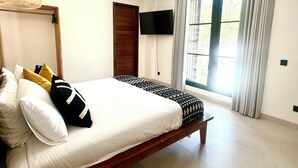 A bedroom with a bed, pillows, a television, a window with curtains, and a hanging light fixture.