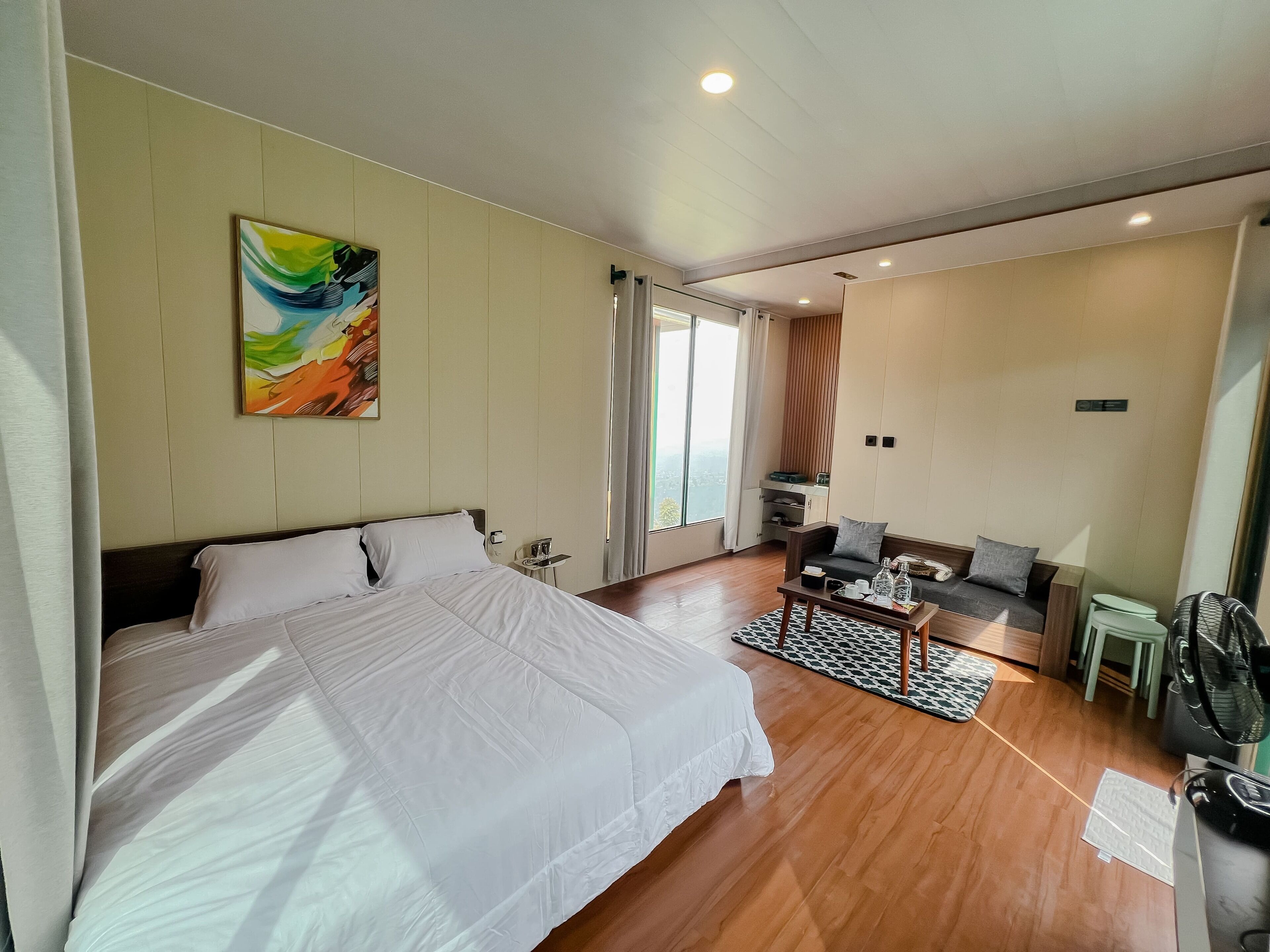 A hotel room with a large bed, a sofa, a small table, and a fan.