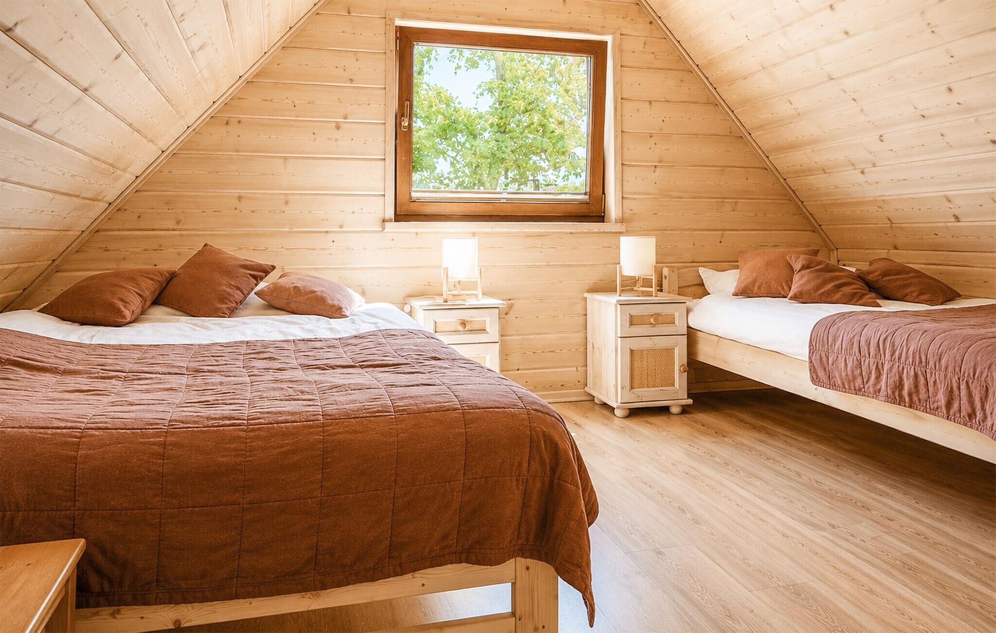 A cozy attic bedroom with two beds, wooden walls, a window, and a nightstand with a lamp.