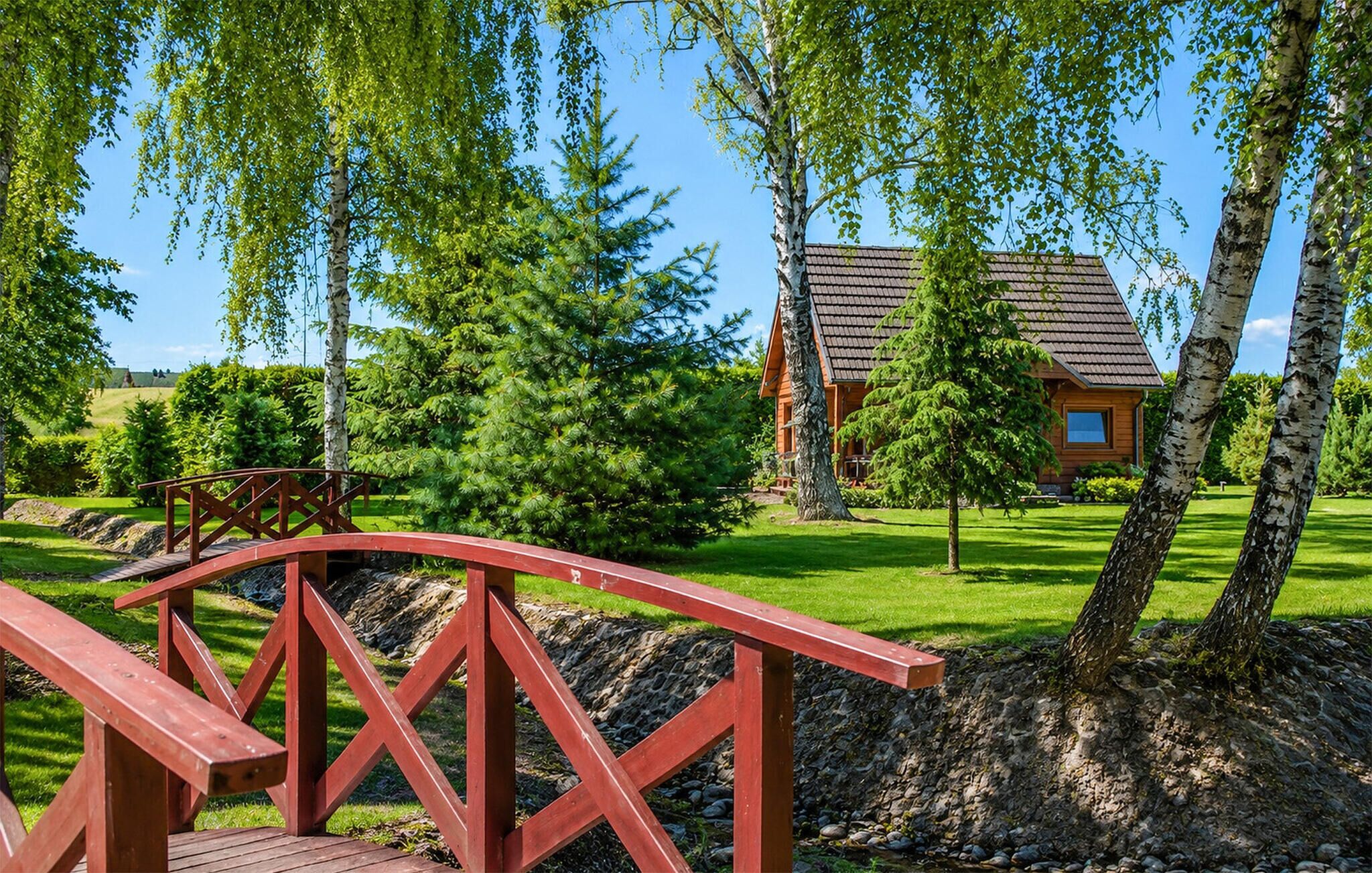 A wooden bridge leading to a cabin surrounded by trees and a grassy area.