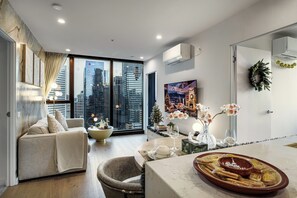 A modern living room with a sofa, a coffee table, and a view of the cityscape through large windows. - Zen Sky Home - Super Centre w/ Carpark & Pool (Melbourne)