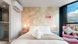 Ein modernes Hotelzimmer mit Wand im Marmoroptik, einem Bett mit weißer Bettwäsche, einem Blick auf die Stadt durch ein großes Fenster und einem rosa Sessel mit floralem Kissen.