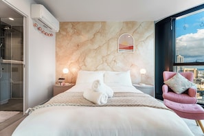 Ein modernes Hotelzimmer mit Wand im Marmoroptik, einem Bett mit weißer Bettwäsche, einem Blick auf die Stadt durch ein großes Fenster und einem rosa Sessel mit floralem Kissen.