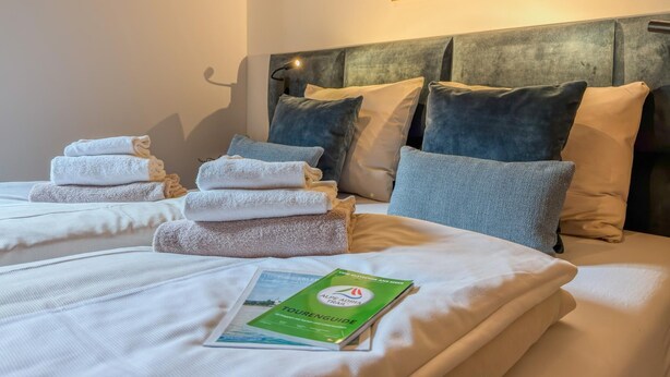A hotel room with a bed, towels, pillows, a headboard, a wall-mounted lamp, and a brochure on the bed. - Modern apartment with Alps and lake views (Afritz am See)