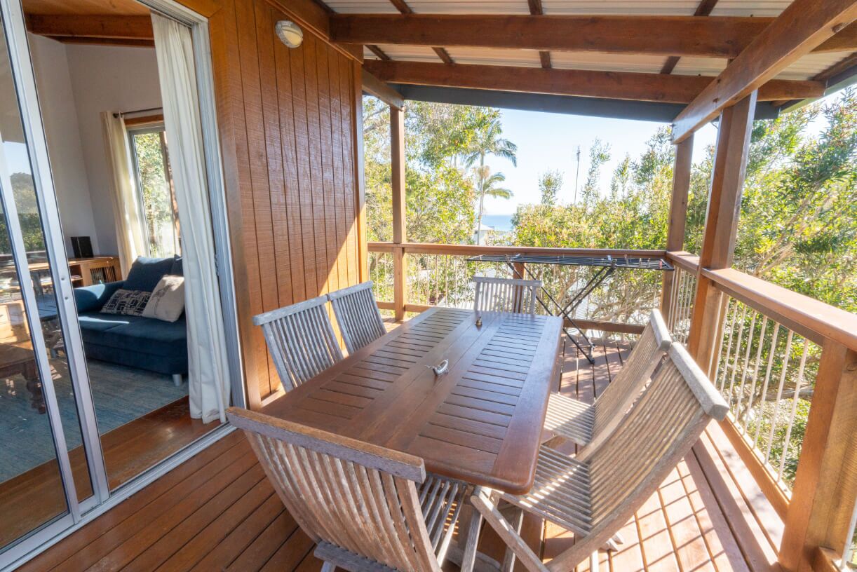 Eine Holzdachterrasse mit Tisch und Stühlen, eine Glastür zu einem Raum mit einer Couch und Blick auf Palmen und das Meer.