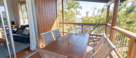 Eine Holzdachterrasse mit Tisch und Stühlen, eine Glastür zu einem Raum mit einer Couch und Blick auf Palmen und das Meer.