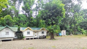Une rangée de petites cabanes à un seul étage, avec un sol de gravier, entourées d’une végétation luxuriante.