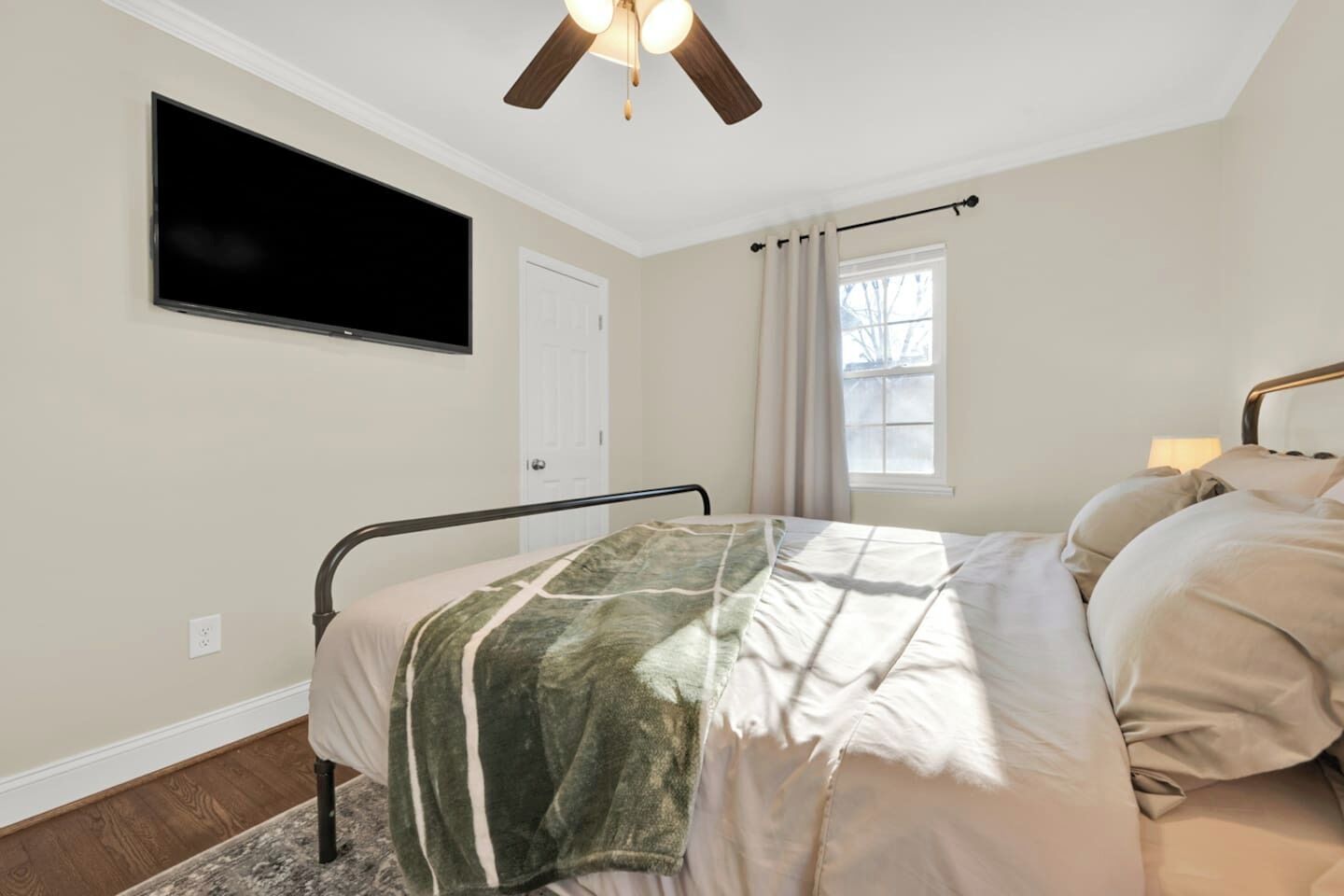 A bedroom with a bed, a ceiling fan, a window with curtains, a wall-mounted TV, and a door.