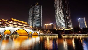 Vista noturna de um horizonte urbano com edifícios modernos altos e uma ponte tradicional com arcos iluminados.