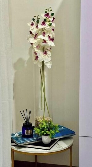 A room with a tall orchid arrangement on a table, a small potted plant, and a book. - The GoodVibes Condotel Red Residences Makati (Makati)