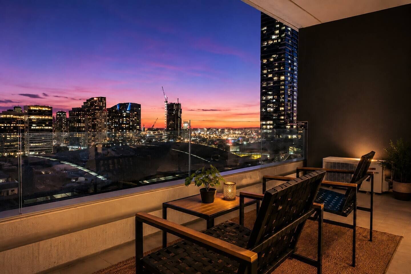 A balcony with a city view at dusk, featuring a table with chairs, a potted plant, and an air conditioning unit.